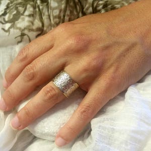 May include: A wide silver ring with an intricate floral pattern is worn on a finger. The ring has a gold-colored band. The ring is a statement piece of jewelry.