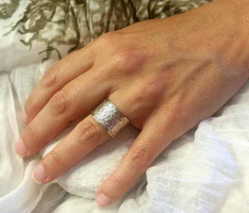 May include: A wide silver ring with an intricate floral pattern is worn on a finger. The ring has a gold-colored band. The ring is a statement piece of jewelry.