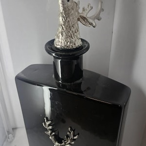 Könnte beinhalten: Eine schwarze, rechteckige Flasche mit einem silbernen Hirschkopf-Verschluss. Der Verschluss zeigt einen detaillierten Hirschkopf mit Geweih. Ein silbernes Emblem eines Hirschgeweihs befindet sich auf der Vorderseite der Flasche. Die Flasche hat eine glänzende Oberfläche.