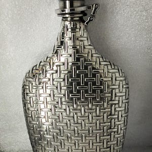 Puede incluir: Una petaca plateada con un diseño de patrón tejido. La petaca tiene una tapa con bisagras y un pequeño tapón redondo. El cuerpo de la petaca es de color plata clara y la tapa es de color plata más oscuro.