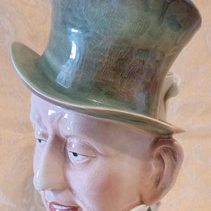 Puede incluir: Una jarra de cerámica con forma de cabeza de hombre que lleva un sombrero de copa verde. El hombre lleva un cuello blanco y una corbata de lazo con estampado rojo y blanco. La jarra tiene un esmalte verde.