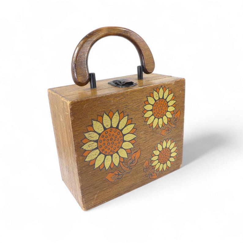 May include: A rectangular wooden handbag featuring a dark brown finish and a rounded wooden handle. The front panel is decorated with three painted sunflowers in shades of yellow and orange, and the top includes a black metal latch closure.