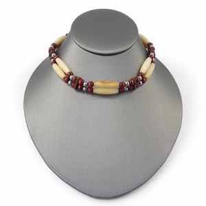 May include: A two-strand necklace on a grey mannequin. It features cream-coloured, cylindrical beads, with small red and silver beads. The central beads are larger and cream-coloured.