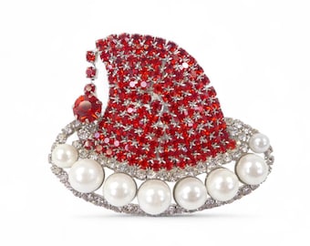 May include: A Christmas-themed brooch shaped like a Santa hat. The hat is adorned with red crystals and edged with a silver-tone border and white pearls. A large red jewel is at the top.