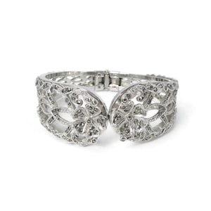 Peut inclure: Un bracelet articulé de couleur argentée présentant un motif floral ajouré complexe. La surface métallique est ornée de nombreuses petites pierres sombres à facettes incrustées dans le filigrane, avec une charnière centrale.