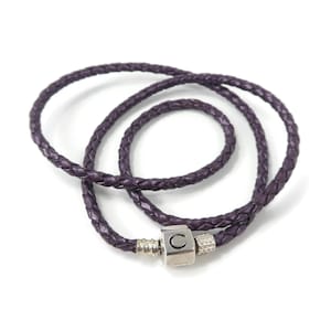 Può includere: Un braccialetto avvolgente in pelle intrecciata viola con chiusura argentata. Il braccialetto è composto da due fili di pelle intrecciata che si avvolgono attorno al polso. La chiusura è di forma quadrata con la lettera "C".