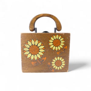 May include: A square wooden handbag featuring a dark brown finish and a semi-circular wooden handle. The front panel is decorated with three painted sunflowers in shades of yellow and orange, each with detailed brown centres and green leaves.