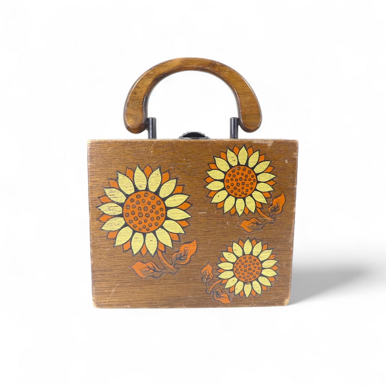 May include: A square wooden handbag featuring a dark brown finish and a semi-circular wooden handle. The front panel is decorated with three painted sunflowers in shades of yellow and orange, each with detailed brown centres and green leaves.