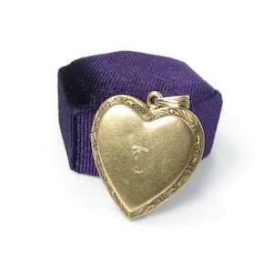 May include: A gold heart-shaped locket featuring an engraved letter C in the centre and intricate scrollwork along the edges. The locket includes a small metal bail at the top for attachment to a chain, showcasing a smooth, polished finish.