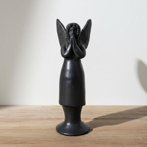 May include: A black angel statue with hands clasped in prayer and wings spread. The figure is made of a dark material and stands on a circular base. The angel's head is bowed, and the wings are detailed.