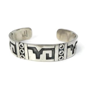 Peut inclure: Un bracelet manchette en argent présentant un motif répétitif de formes géométriques et de colonnes verticales de trois petits cercles. L'intérieur de la bande est estampillé du texte MEXICO et d'autres marques, avec une finition métallique polie.