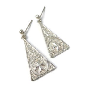 May include: A pair of silver filigree earrings. Each earring has a triangular shape with a star design in the centre. The earrings are dangling and have a small ball at the top for the ear wire. The filigree work is intricate and detailed.