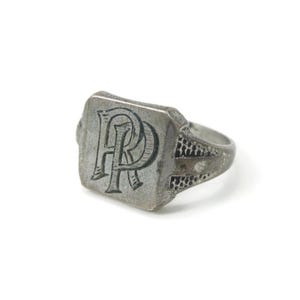 May include: A silver ring with a rectangular face. The face displays the intertwined letters "R" and "P" in a decorative font. The band has a textured pattern on the sides. The ring is likely made of metal and has an aged appearance.