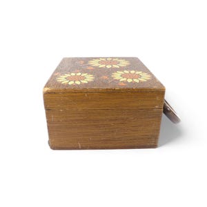 May include: A rectangular wooden box with a hinged lid featuring a painted design of three yellow and orange sunflowers. The box has a natural brown wood grain finish and a small metal ring pull attached to the side for opening the lid.