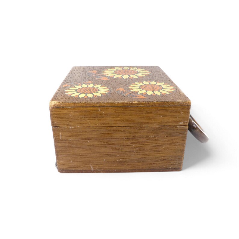 May include: A rectangular wooden box with a hinged lid featuring a painted design of three yellow and orange sunflowers. The box has a natural brown wood grain finish and a small metal ring pull attached to the side for opening the lid.