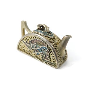 May include: A small silver teapot with detailed filigree and enamel work. The teapot has a curved handle, a spout, and a lid with blue and gold accents. The body of the teapot is decorated with floral patterns.