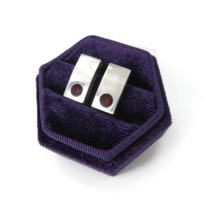 May include: A pair of rectangular silver-toned cufflinks, each featuring a single circular dark red stone inlay. The cufflinks are presented inside a hexagonal purple velvet jewellery box with a soft, textured fabric lining and raised edges.