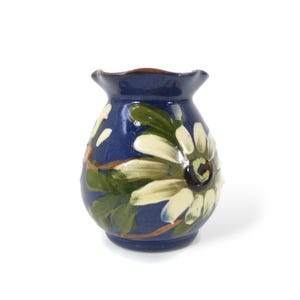 May include: A small, hand-painted ceramic vase with a dark blue background. The vase features a large, cream-coloured flower with green leaves. The rim of the vase is a darker brown colour. The vase is a decorative item.