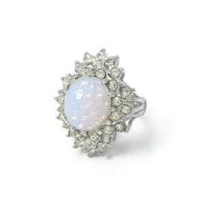 Könnte beinhalten: Ein silberfarbener Ring mit einem großen, ovalen, schillernden weißen Cabochon-Stein in der Mitte. Der Stein ist von einem Heiligenschein aus kleinen, klaren, rund geschliffenen Kristallen umgeben, die in einem gewellten, sternförmigen Metallrahmen sitzen.