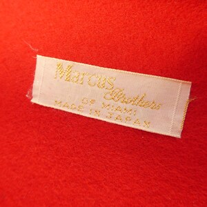 May include: A bright red fabric surface featuring a rectangular white woven label with gold embroidered text that reads Marcus Brothers of Miami, Made in Japan. The label is stitched onto the vibrant red textile material.