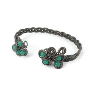 Puede incluir: Un brazalete tipo puño hecho de alambre oscuro y oxidado trenzado en una banda. Los extremos abiertos presentan intrincadas espirales y bucles de alambre, cada uno acentuado con pequeñas cuentas verdes, redondas y lisas. Diseño simétrico.