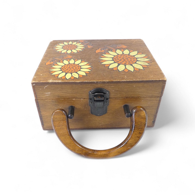 May include: A rectangular wooden box with a hinged lid featuring painted yellow and orange sunflower designs. The box includes a dark metal latch closure on the front and a curved, polished wooden handle attached to the front panel.