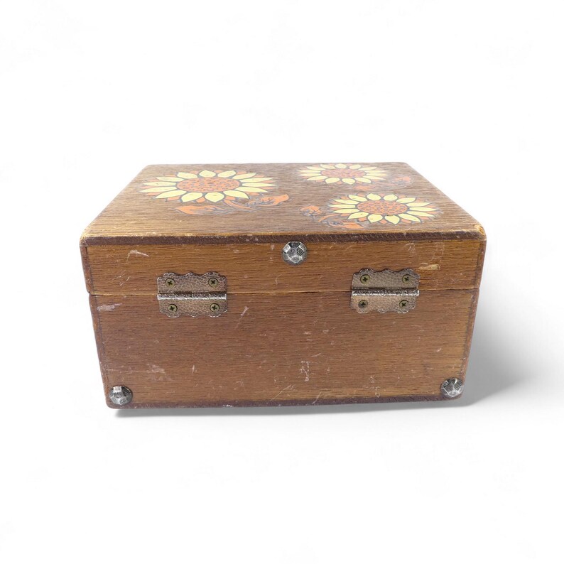 May include: A rectangular wooden box with a natural brown finish, featuring a painted sunflower design on the lid. The box includes two metal hinges on the back, a small metal latch on the front, and decorative metal studs on the bottom corners.