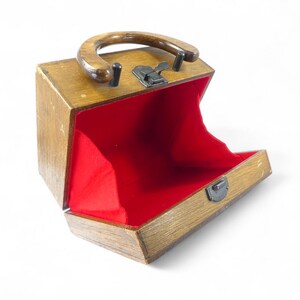 May include: A wooden box-style handbag with a polished brown finish, featuring a curved wooden handle, a metal latch closure, and a vibrant red fabric interior lining. The structure is rectangular with a hinged opening that reveals the interior.