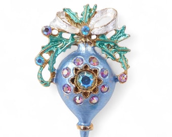 May include: A blue Christmas ornament brooch with a gold-coloured trim. The brooch features a white bow, teal leaves, and several iridescent rhinestones. The ornament has a central, large, blue rhinestone.