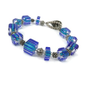 Può includere: Un bracciale con perle di vetro cubiche a righe blu e turchesi traslucide, alternate a piccoli distanziatori in metallo argentato e perle rotonde trasparenti. Il gioiello è fissato con una chiusura a T di colore argentato.