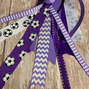 May include: Purple and white hair tie with soccer ball ribbon. The hair tie has a purple elastic band and multiple ribbons with white and purple chevron patterns, solid purple, and soccer ball designs.