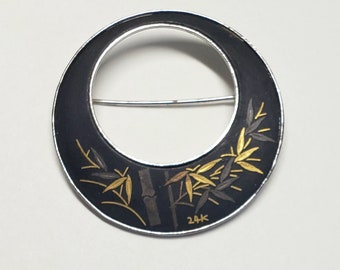 Può includere: Una spilla circolare in argento con sfondo in smalto nero e un design di bambù in oro e argento. La spilla ha una spilla sul retro e l'iscrizione "24K" sul fondo.