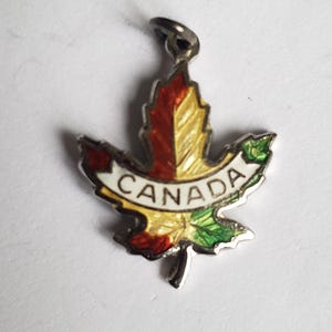 May include: A silver-toned maple leaf charm with a colorful enamel design. The leaf is divided into sections of red, yellow, and green. A white banner across the center reads "CANADA". The charm has a small loop for attaching to a chain or bracelet.