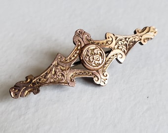 May include: Ornate gold-toned brooch with intricate floral and scrollwork details. The brooch has a central circular element with a floral design and a pin closure on the back. The design is symmetrical and features a vintage aesthetic.