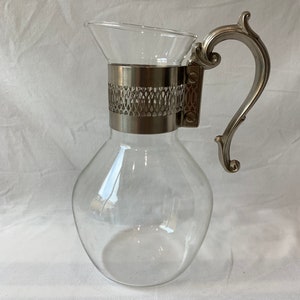 May include: A clear glass pitcher with a bulbous body and a flared top. It features a decorative silver-toned metal band and handle. The handle has an ornate, curved design. The pitcher is empty and sits on a white surface.
