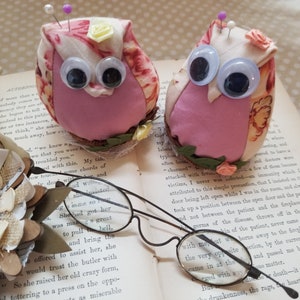 May include: Two owl-shaped pincushions with googly eyes, floral fabric, and pins, resting on an open book. Vintage spectacles and paper flowers complete the scene. A delightful sewing accessory.