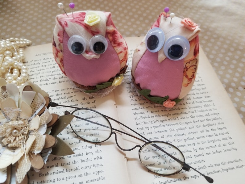 May include: Two owl-shaped pincushions with googly eyes, floral fabric, and pins, resting on an open book. Vintage spectacles and paper flowers complete the scene. A delightful sewing accessory.