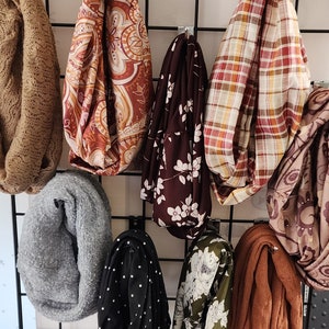 May include: Assortment of eight infinity scarves in various colors and patterns, including brown, gray, black, white, red, and green. The scarves are made of different fabrics, including knit, woven, and printed.