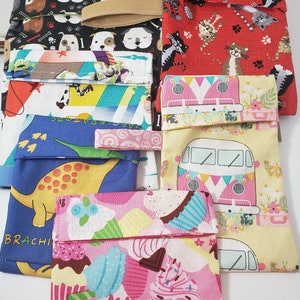 May include: A selection of patterned fabric pouches featuring designs such as dogs, cats, cupcakes, dinosaurs, and a camper van. Each pouch has a fold-over flap and a matching elastic band or strap.