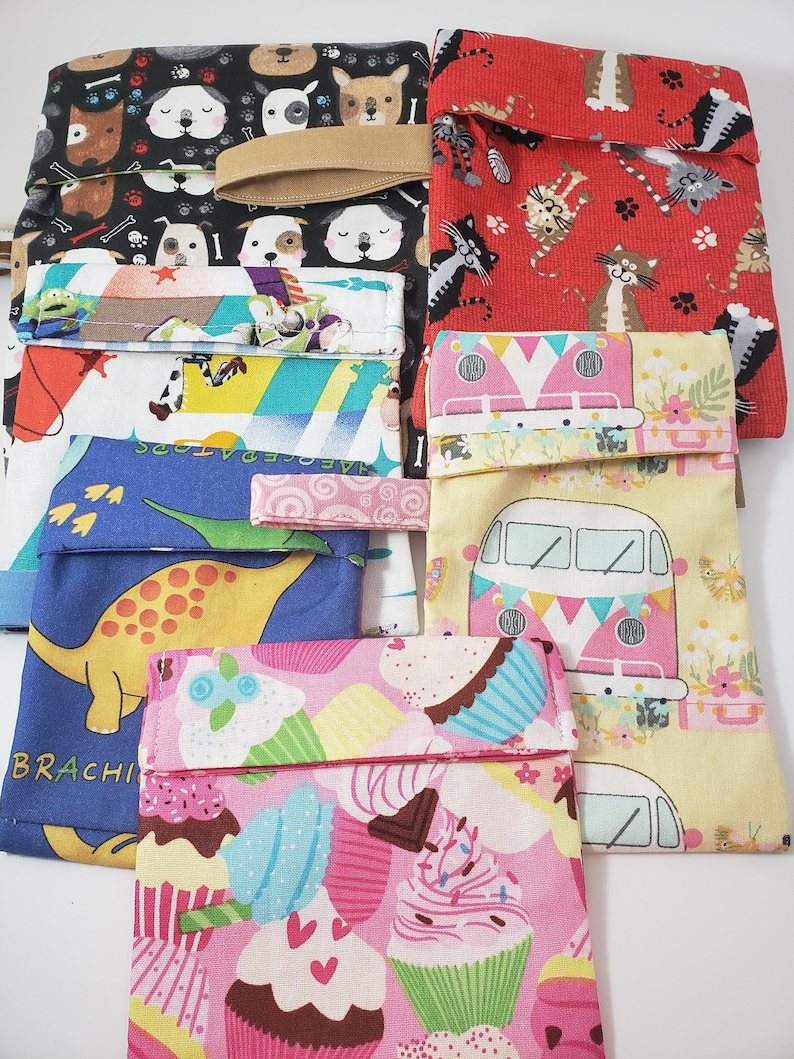 May include: A selection of patterned fabric pouches featuring designs such as dogs, cats, cupcakes, dinosaurs, and a camper van. Each pouch has a fold-over flap and a matching elastic band or strap.