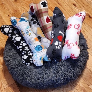 May include: Six plush dog toys shaped like bones in a gray dog bed. The toys are made of soft fabric and have different patterns, including paw prints, hearts, and plaid.