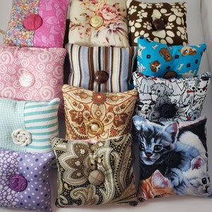 May include: A selection of small, square pin cushions with diverse fabric patterns and button details. Designs include floral, paisley, stripes, polka dots, and animal prints. Each cushion is approximately 7.6 cm square.
