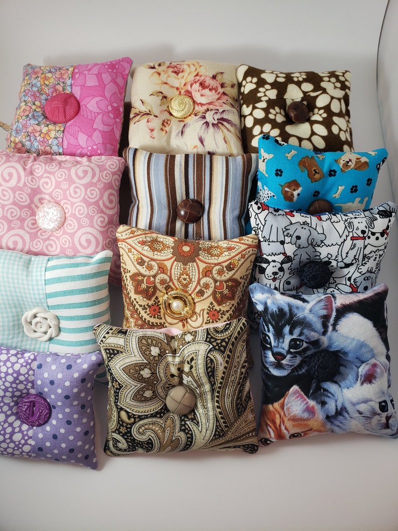 May include: A selection of small, square pin cushions with diverse fabric patterns and button details. Designs include floral, paisley, stripes, polka dots, and animal prints. Each cushion is approximately 7.6 cm square.