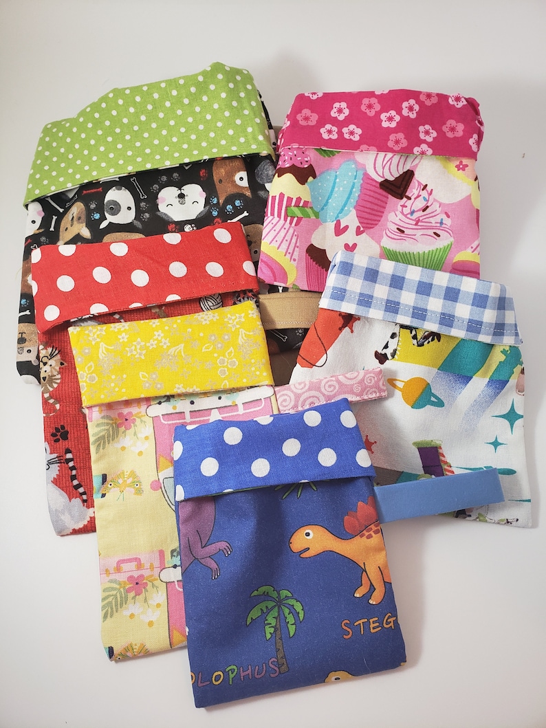 May include: A selection of patterned fabric pouches. Designs include polka dots, cupcakes, dogs, and dinosaurs. Each pouch has a contrasting, folded-over top. Some have a small fabric tab.