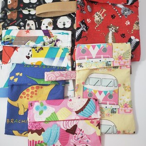 May include: A selection of fabric pouches featuring diverse patterns. Designs include dogs, cats, cupcakes, dinosaurs, and a retro van. The pouches are in various colours, including black, red, pink, yellow, and blue. Some pouches have a fabric loop.