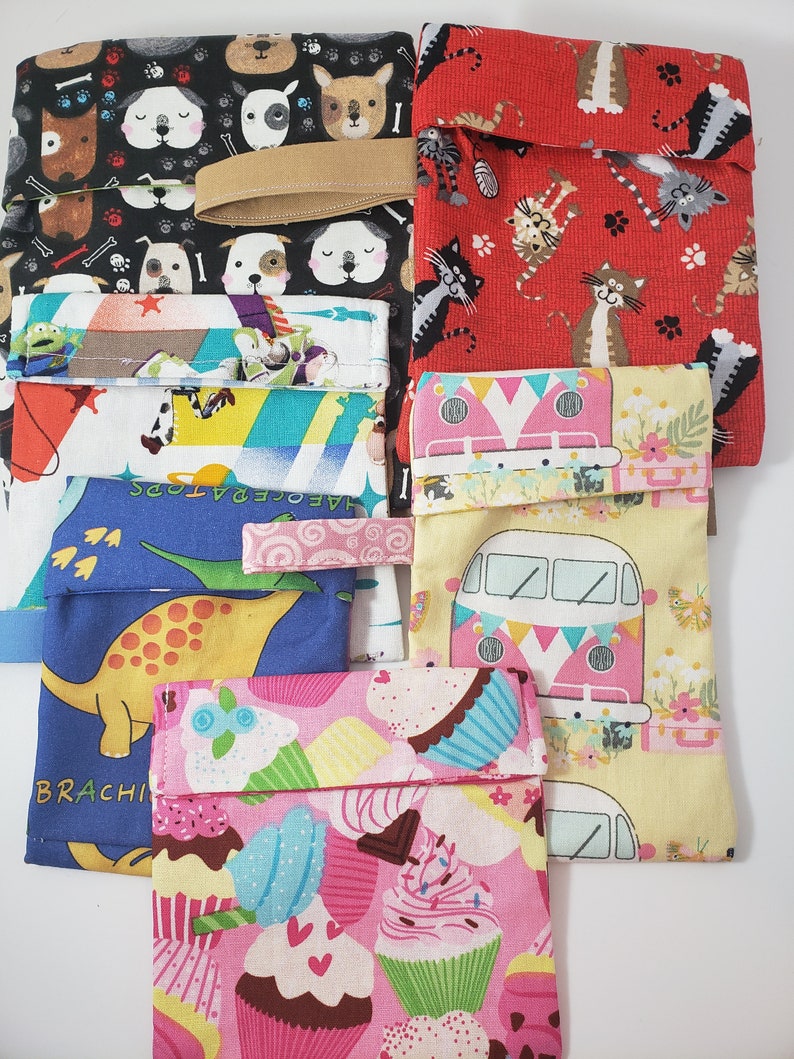 May include: A selection of fabric pouches featuring diverse patterns. Designs include dogs, cats, cupcakes, dinosaurs, and a retro van. The pouches are in various colours, including black, red, pink, yellow, and blue. Some pouches have a fabric loop.