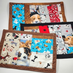 May include: Two brown fabric placemats with a patchwork design featuring various dog breeds. The placemats have a blue, red, and white color scheme with the text "Woof I Woof You" and "Club" on them.