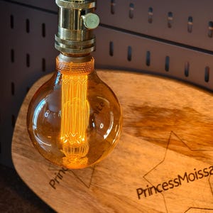May include: A decorative light bulb with a warm orange glow, suspended from a brass fixture. The bulb is positioned above a wooden surface with the text "PrincessMoissanite" and a star design etched into it. The background features a dark metal panel.