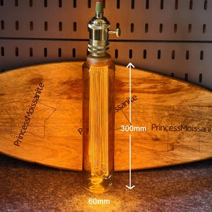 May include: A vintage-style light fixture with a clear glass bulb, 300mm in length, and a brass base. The bulb is filled with vertical filaments. The fixture is suspended by a red cord. The fixture is placed in front of a wooden board with the text "PrincessMoissanite".