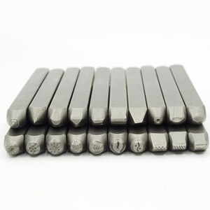 May include: A set of 20 metal letter stamps with various designs, including letters, numbers, and symbols. The stamps are arranged in two rows, with the top row slightly offset from the bottom row. The stamps are all silver in color and have a polished finish.